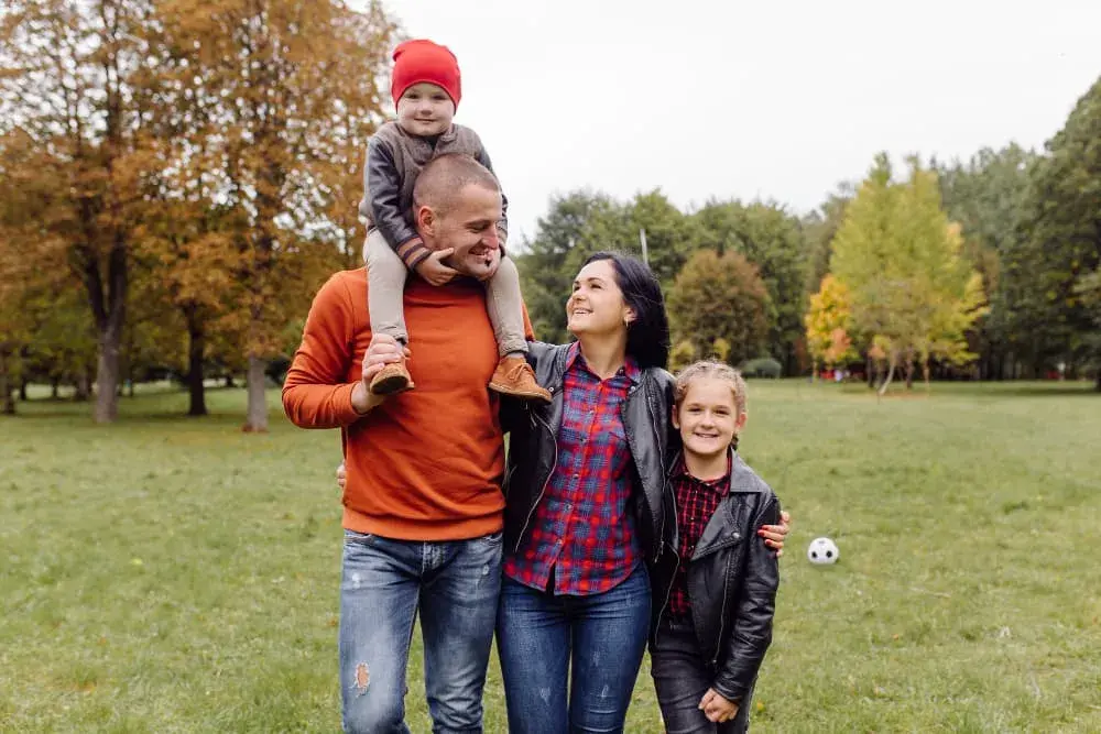 A happy family enjoys time together in a park on a cool day. The father carries a young child on his shoulders while the mother and older child walk beside them, symbolizing joy, stability, and the financial freedom that comes with smart refinancing through Chris Lewis Home Loans.