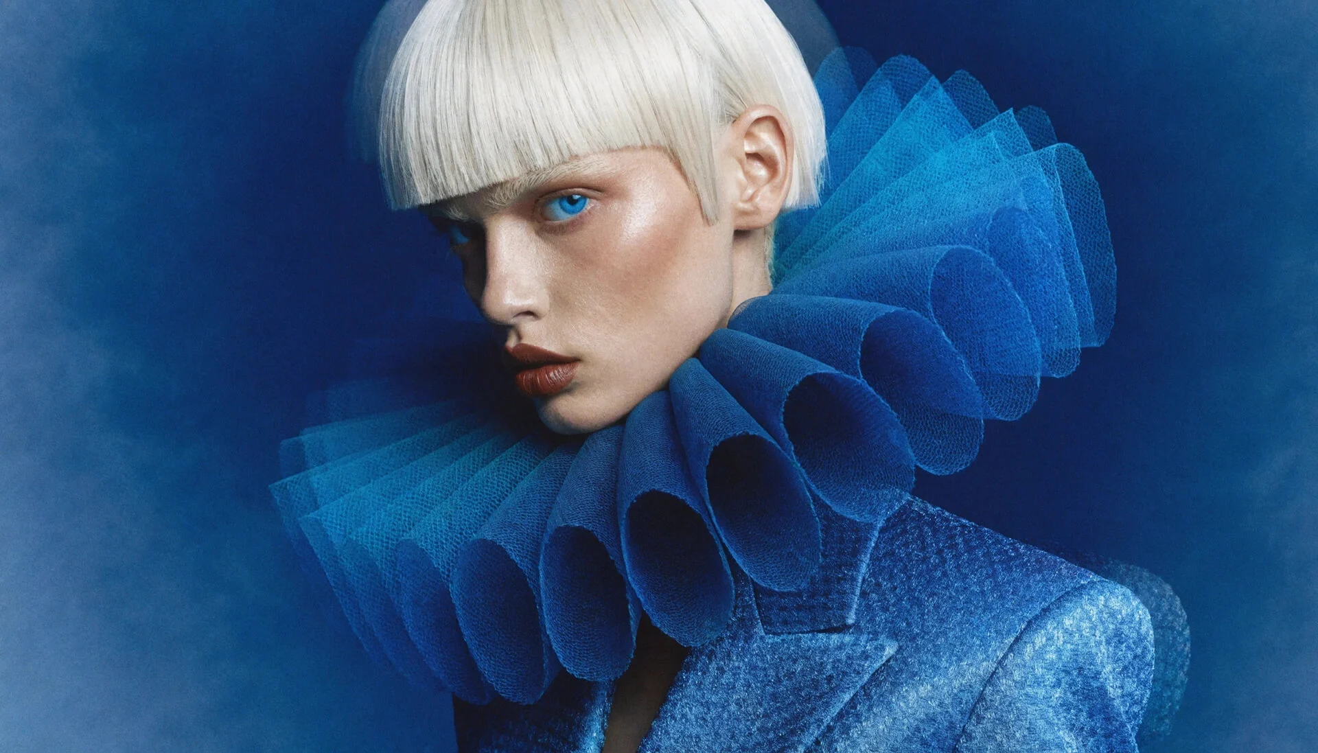 Close-up fashion portrait of a model with short platinum hair wearing a vivid blue outfit with a sculptural ruffled collar