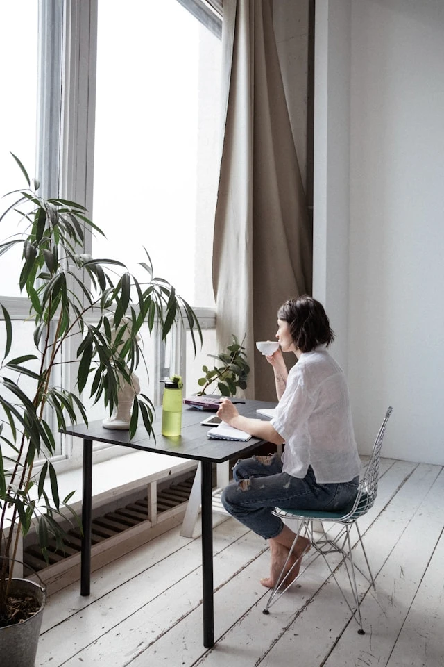 Woman sitting by a window next to a plant, reflecting calmly and focusing on long-term progress.