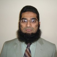 Elderly man with a gray beard smiles warmly while wearing a dark suit with a blue tie. The background is a neutral gray, conveying a professional tone.