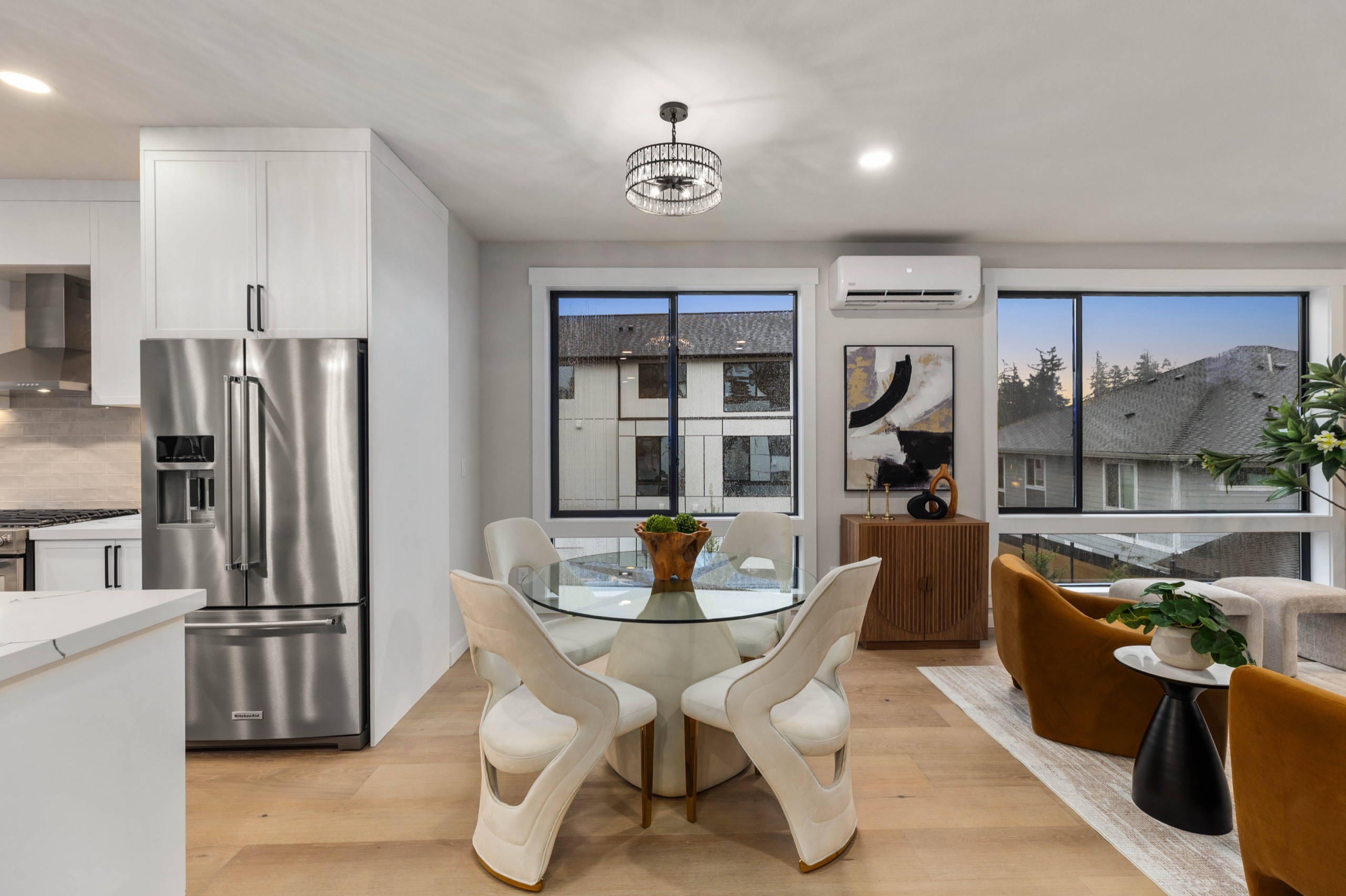 Elegant luxury dining nook with round glass table, sculpted white chairs, floor-to-ceiling windows, and open view to modern kitchen with stainless steel refrigerator