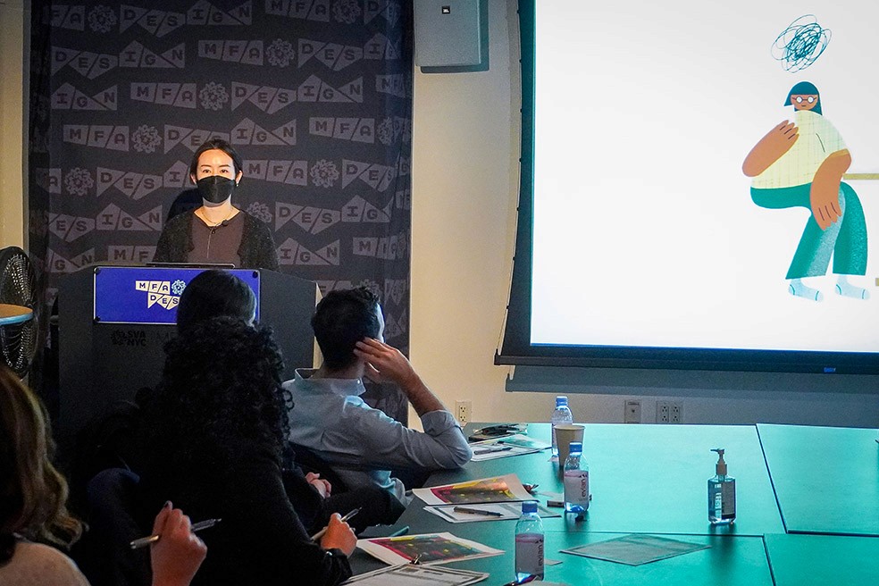 A woman dressed in brown and black is giving a speech to the audience and an image is projected on the right side of the wall with an illustration of a woman sitting on a chair.
