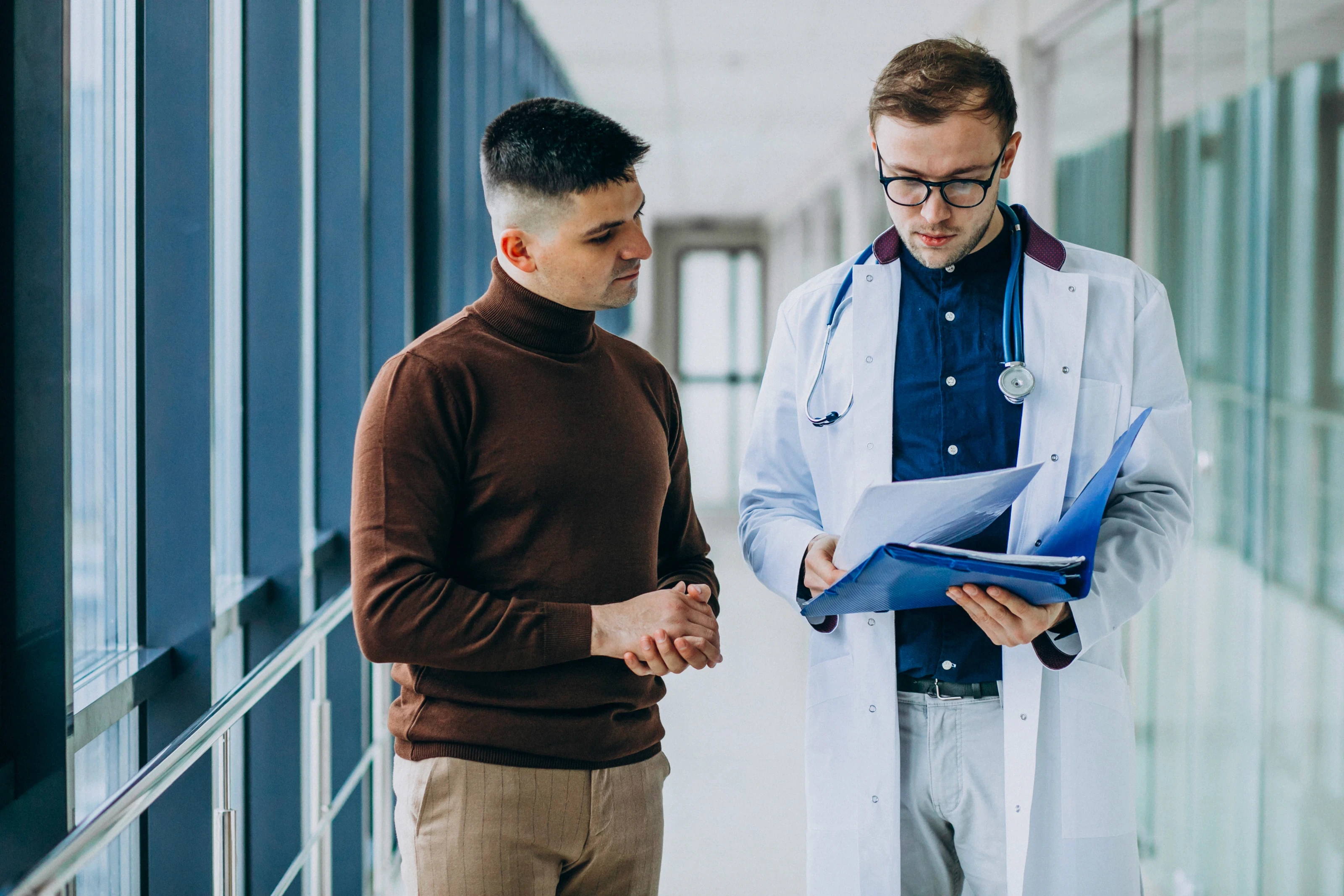 A doctor and patient converse in a bright hospital hallway. The doctor, in a white coat, reviews documents while the patient listens attentively.