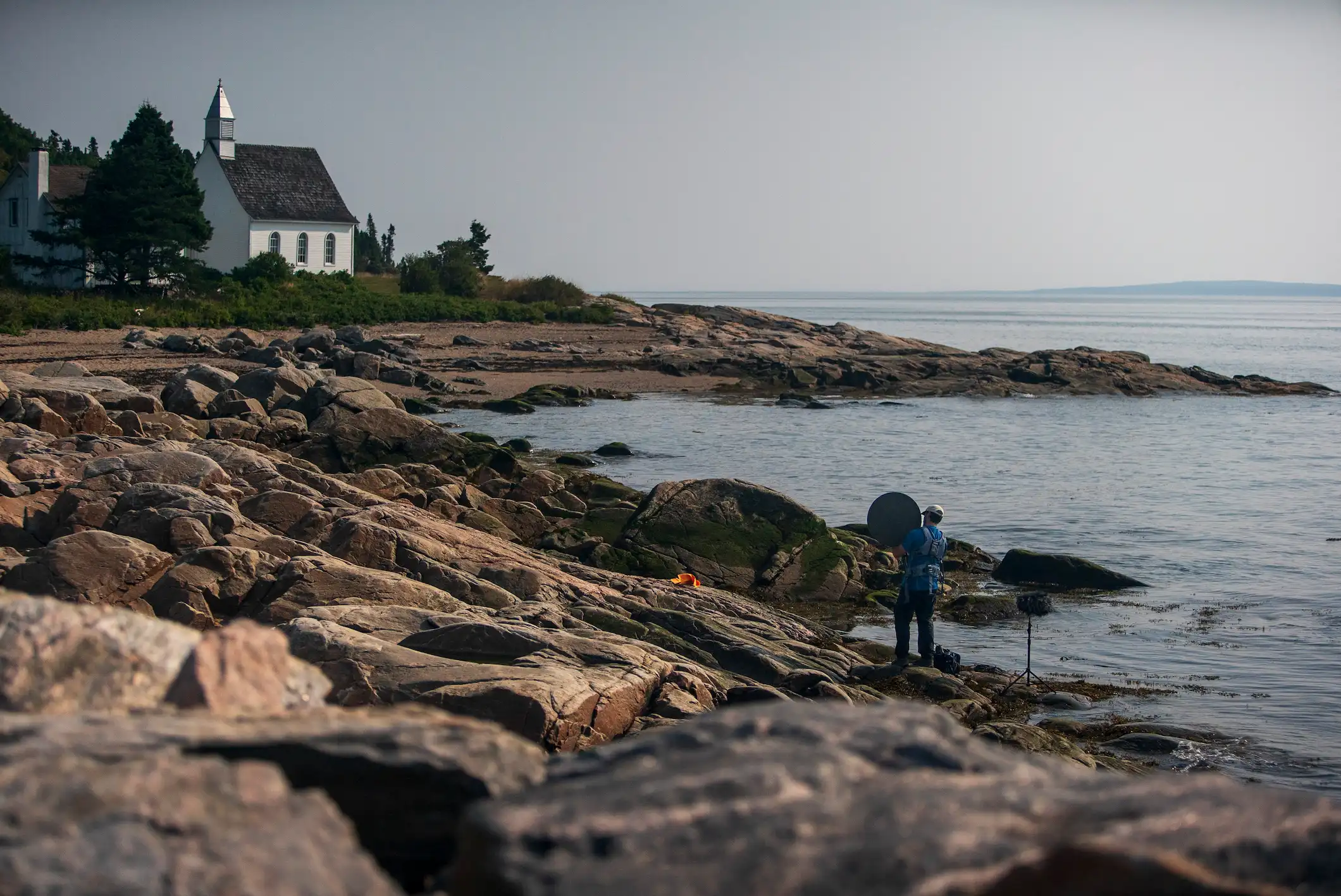 Personne installant du matériel audio sur des rochers au bord de la mer, avec une petite église blanche visible à l’arrière-plan.