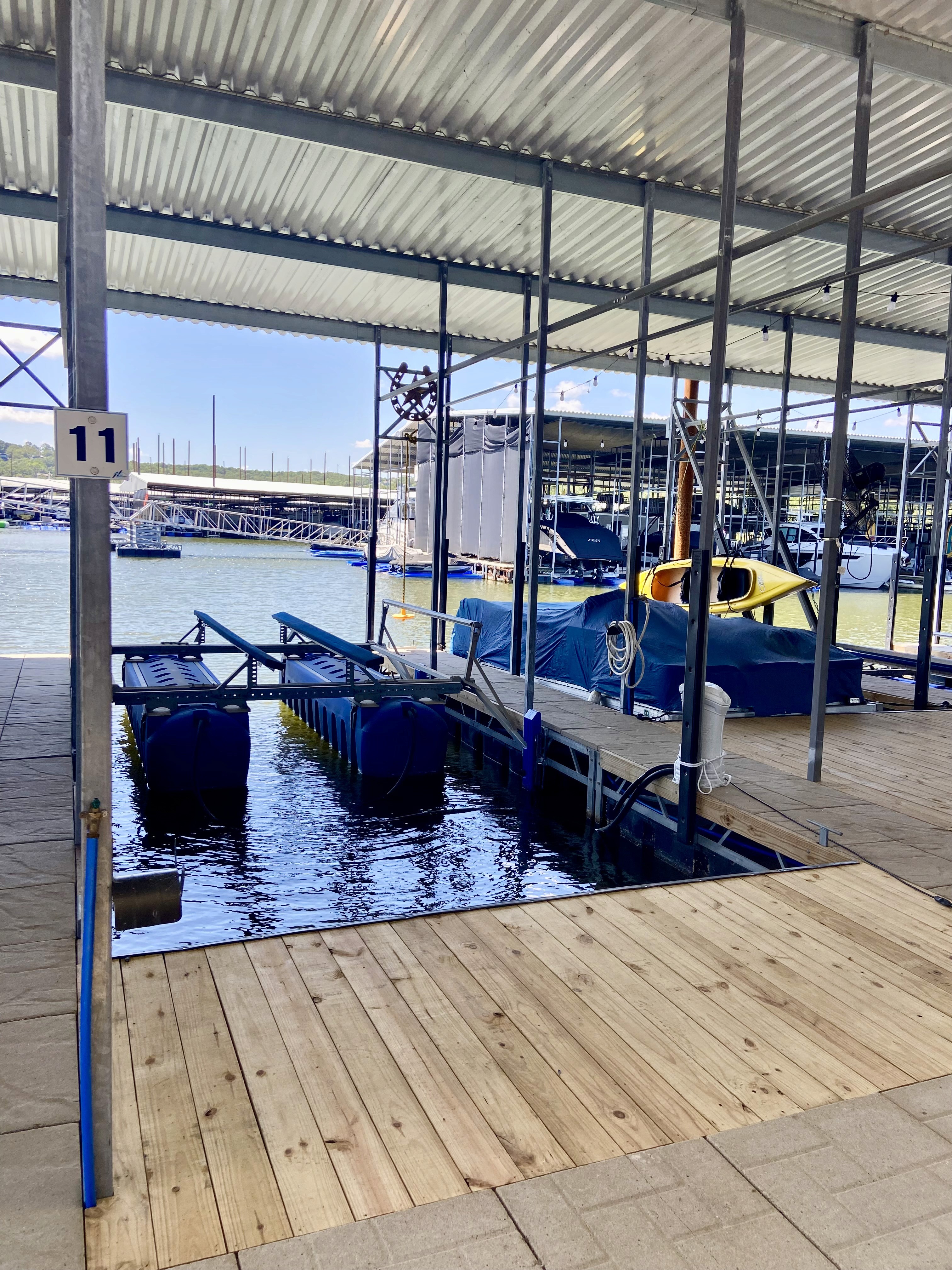 A modern boat dock with wooden decking leads to moored boats under a metal canopy, set against a backdrop of a calm marina and clear blue sky.