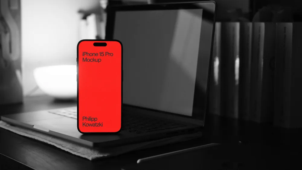 Mockup showcasing an iPhone 15 Pro with a vibrant red screen, placed in front of an open laptop on a desk. The background features a blurred black-and-white setup with books and a bowl for contrast.