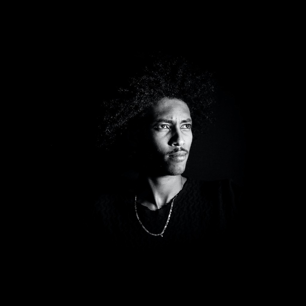 Black white portrait showing a man with curly hair,  black tshirt and a neckles looking away from the camera 