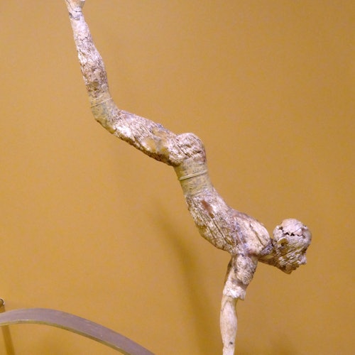 Ancient balancing sculpture of a person performing a handstand, mounted on a curved metallic stand against a beige background.