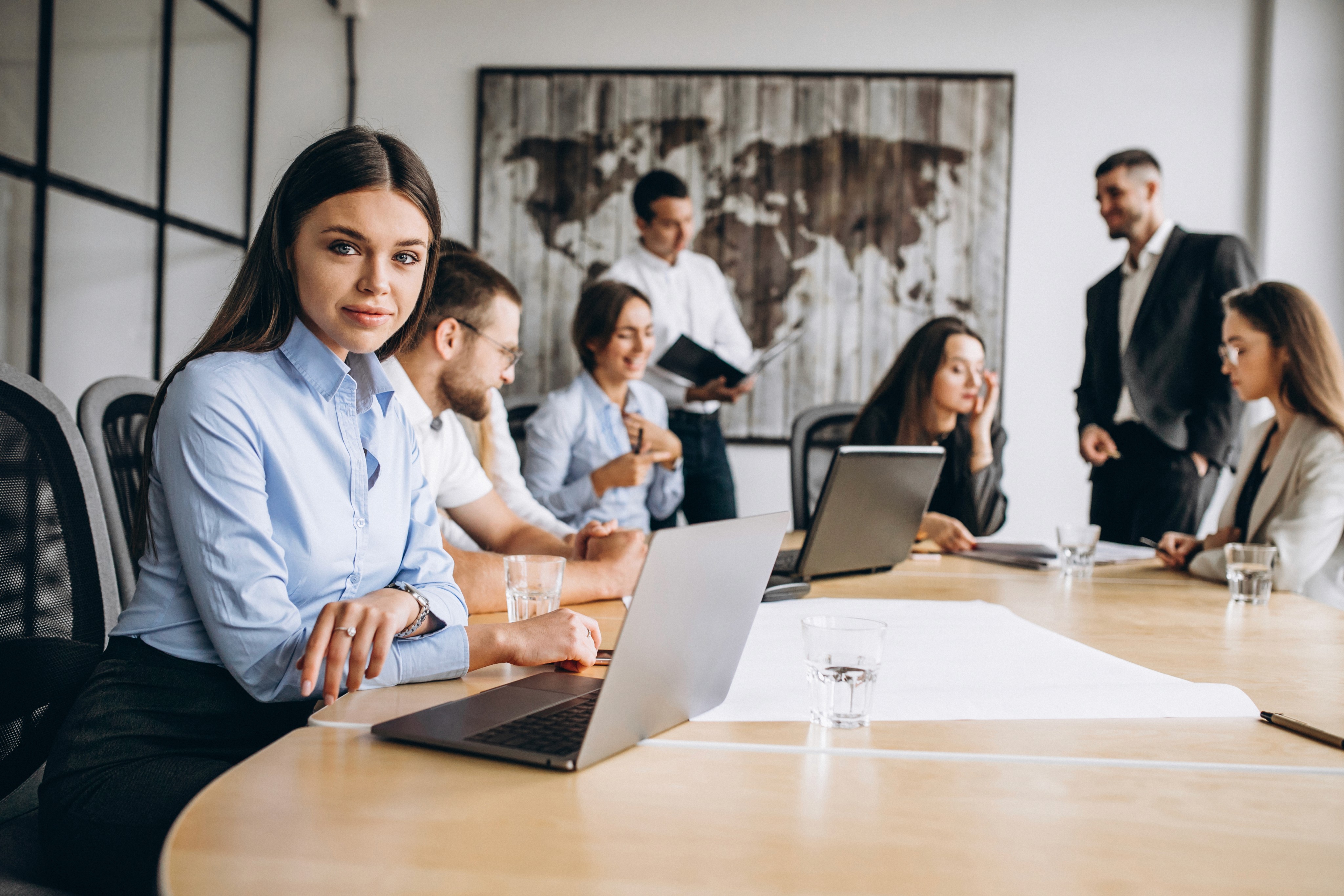 A diverse group of businesspeople in a meeting room with laptops and notebooks, one woman in a blue shirt looking at the camera.