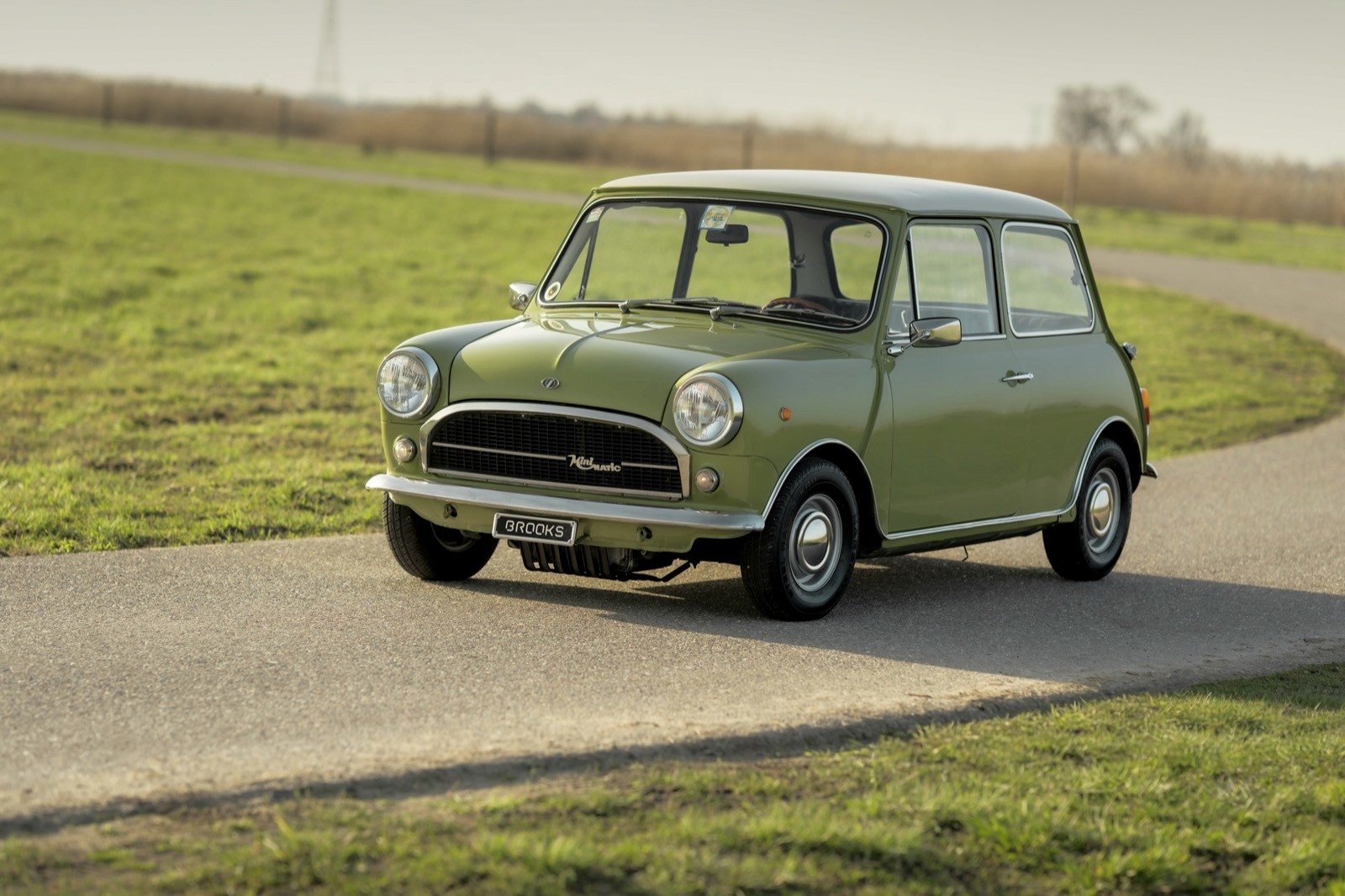 A classic green car drives along a winding road, surrounded by grassy fields under a clear sky.