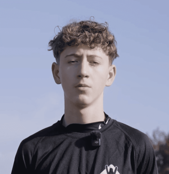Person in floral shirt with glasses standing outdoors near wooden shed and yellow vehicle.