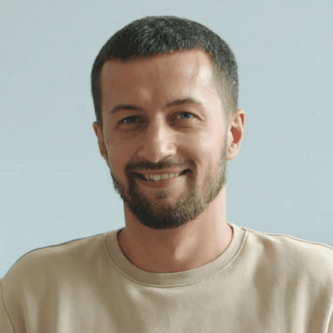 A circular headshot of a smiling man with short, dark hair and a groomed beard, wearing a neutral tan crewneck sweatshirt against a plain gray background.