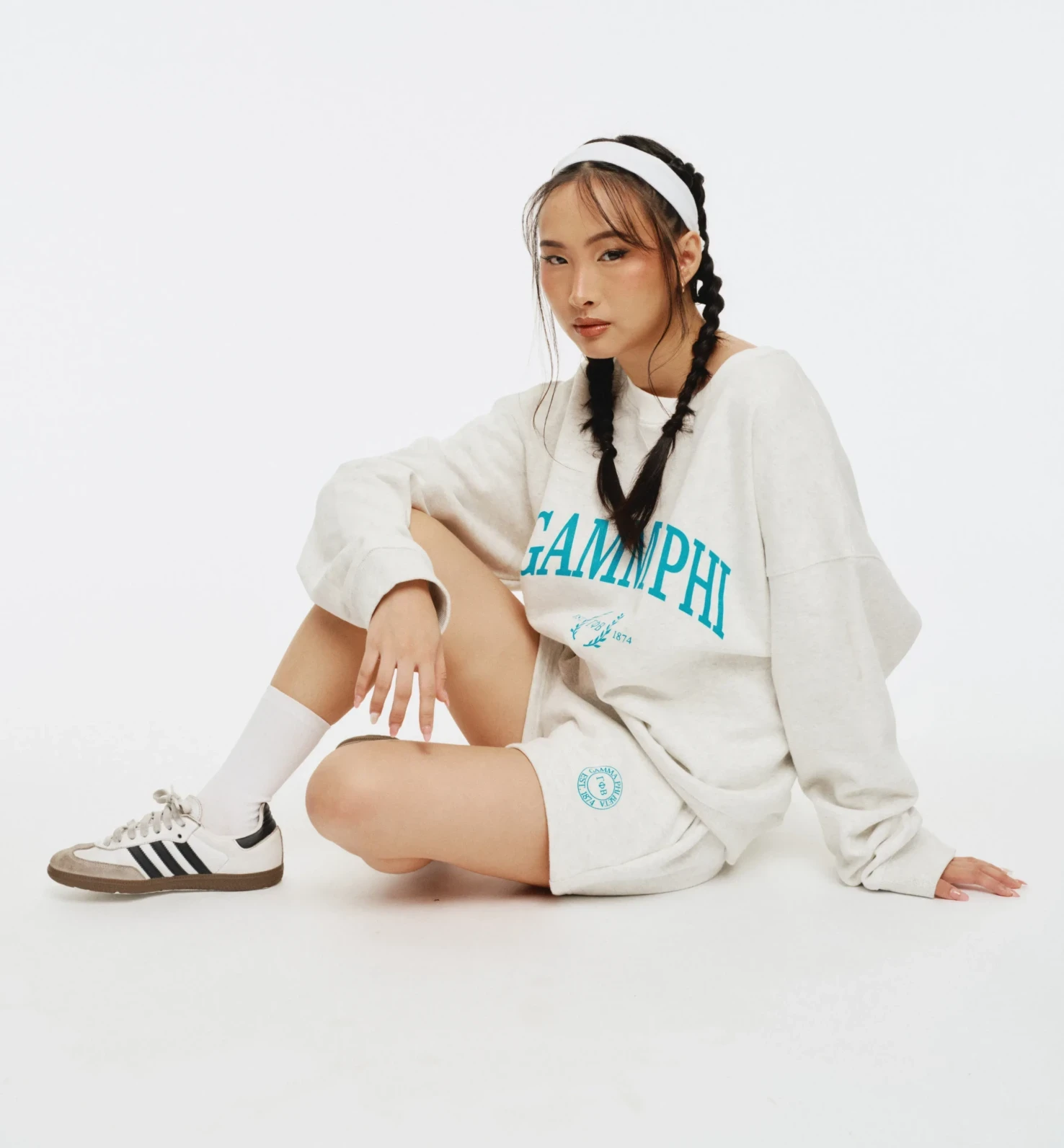 Model sitting on white backdrop wearing a cream “GAMMA PHI” sweatshirt with teal lettering and matching shorts with a small circular crest.
