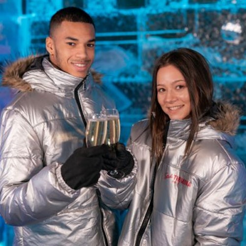 Nasmiješeni par u srebrnim parkama drži čaše šampanjca, stojeći ispred ledenog zida.