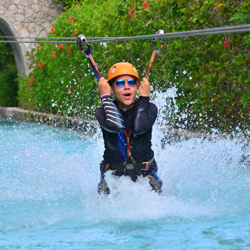 Persona en una tirolesa salpicando en una piscina, usando un casco y gafas de sol, con vegetación y un arco de piedra de fondo.