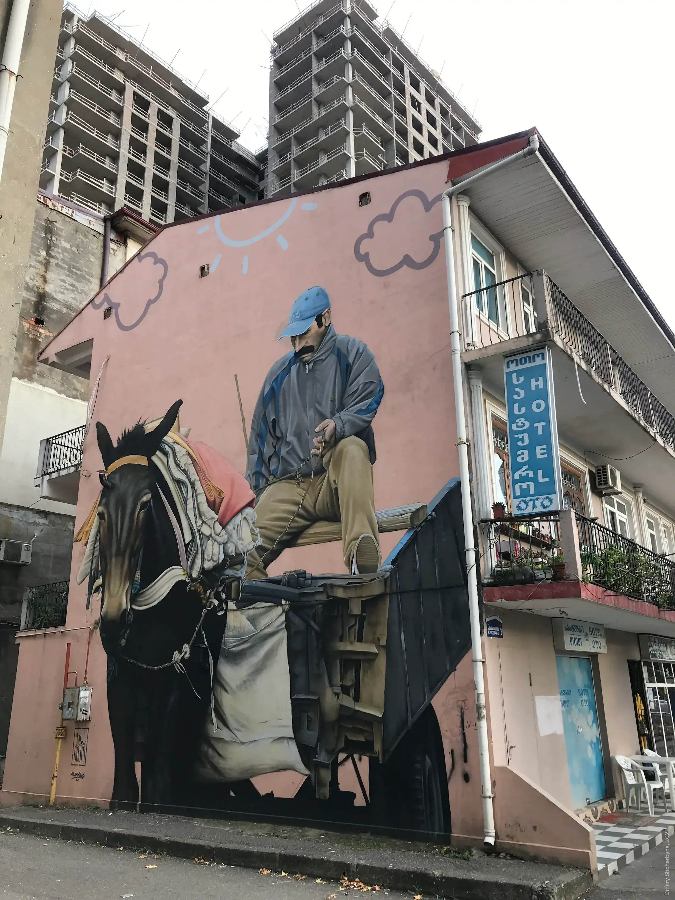 A mural on the side of a small building shows a realistic depiction of a man in a blue jacket and cap sitting on a wooden horse-drawn cart with a dark horse pulling it along in a harness.