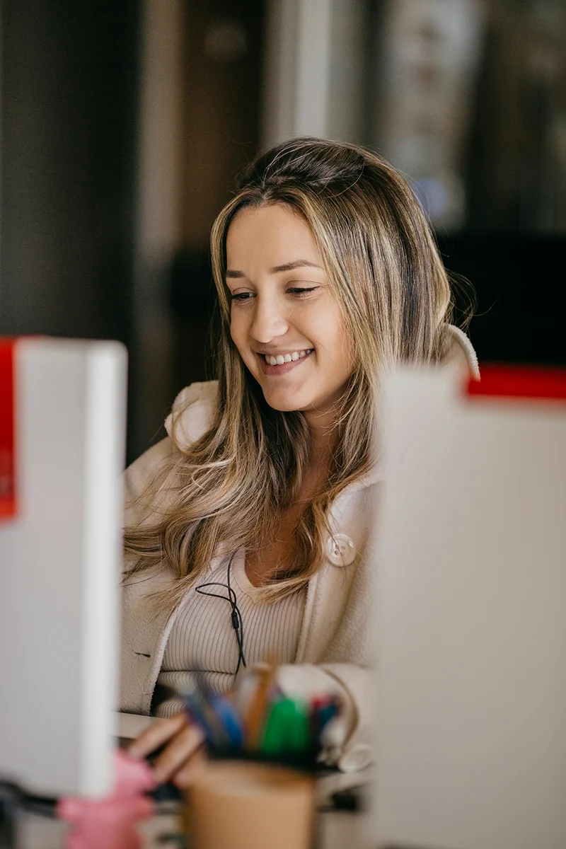 A close-up portrait of a smiling young woman with highlighted hair looking down at a screen or document in a bright setting.
