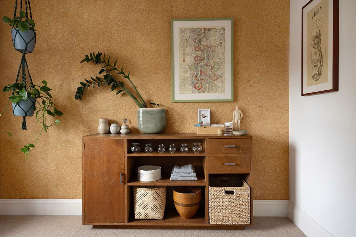 A homely interior view of the Fen Acupuncture clinic showing a warm cork-lined wall, a wooden sideboard with treatment supplies, and indoor plants.