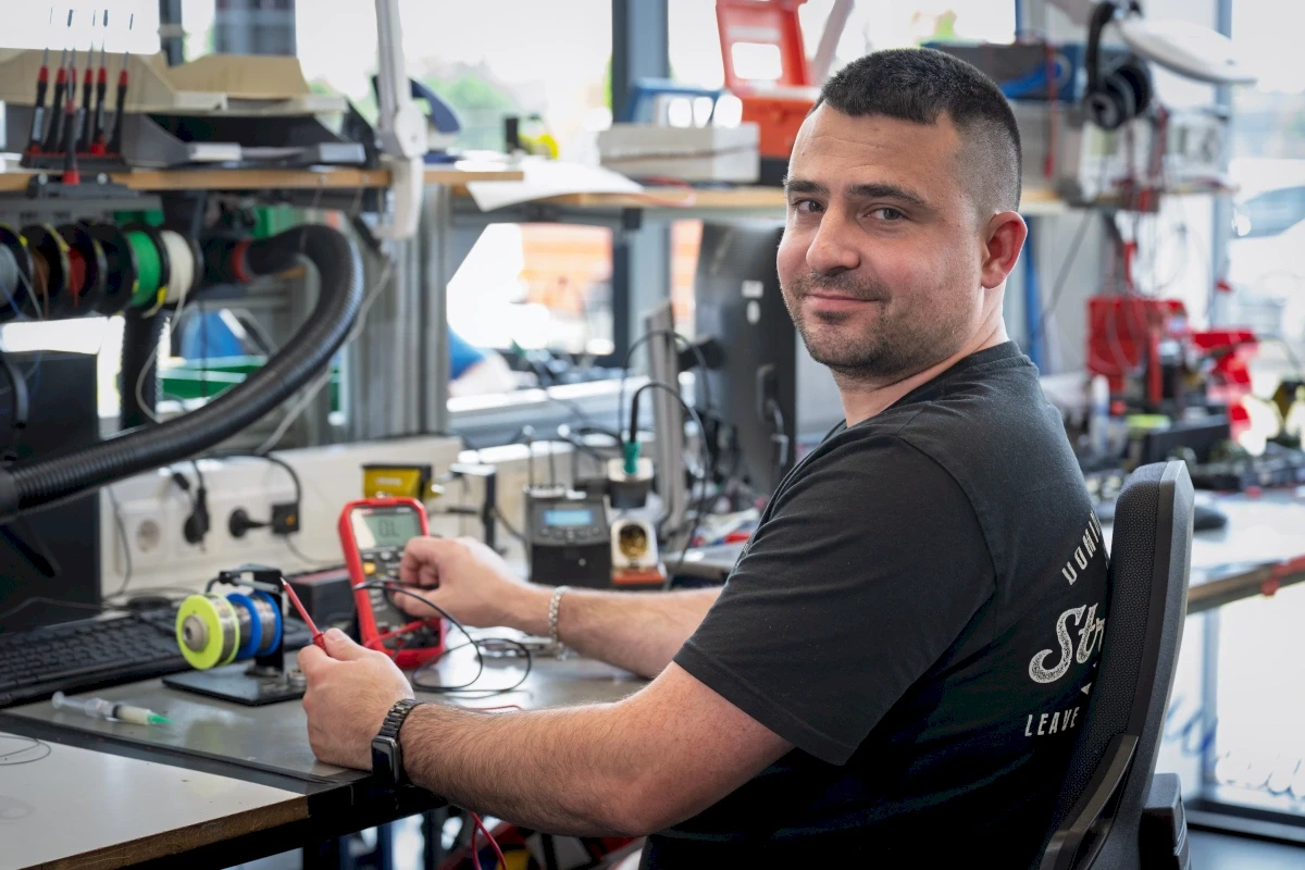 Man using a multimeter to test electronics at a desk in a workshop. He is smiling while working.