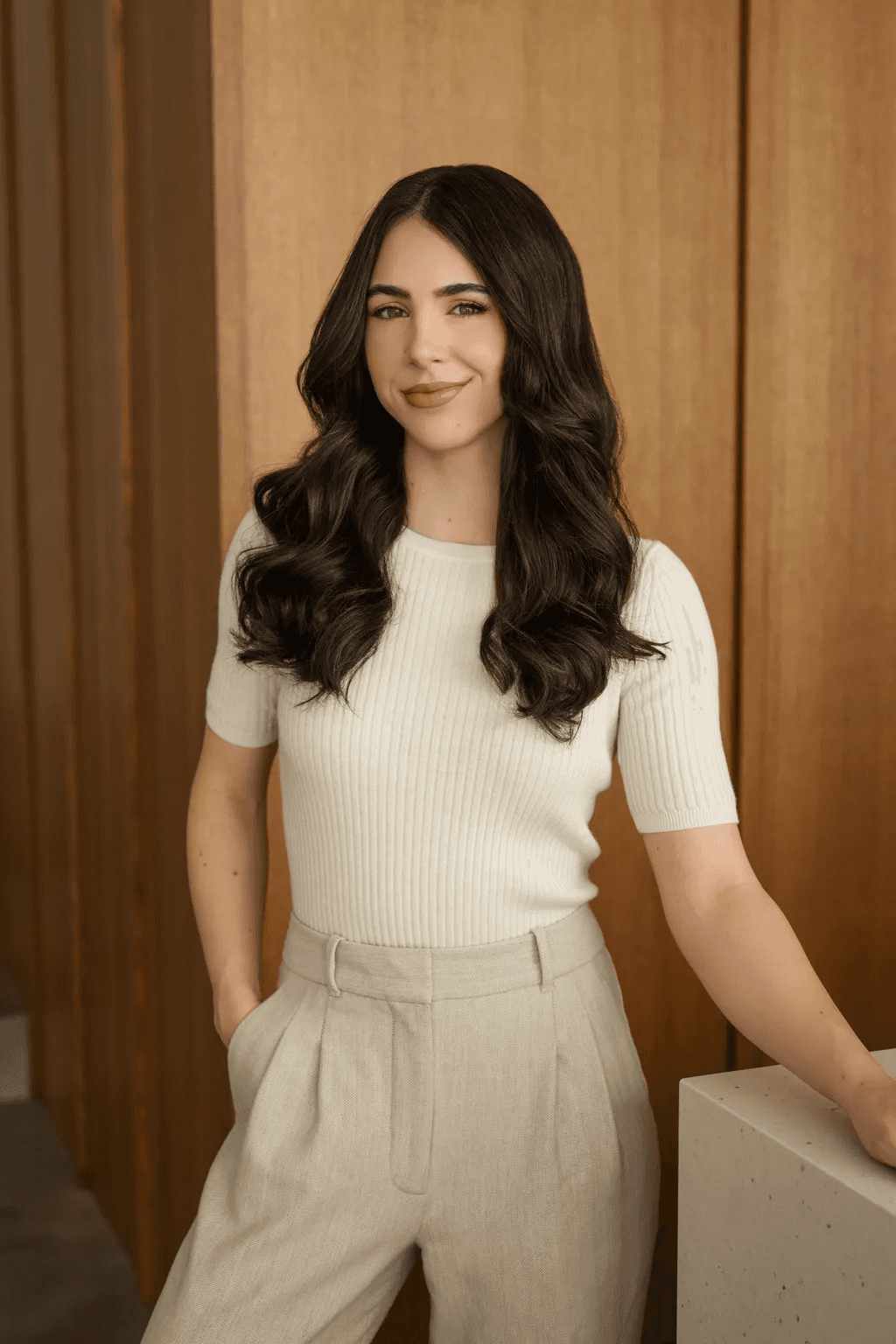 A person with long dark hair and wearing a white ribbed short-sleeve sweater and beige high-waisted trousers stands confidently against a wooden paneled wall.