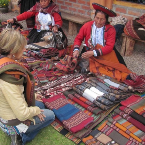 兩位穿著傳統服裝的女性在地上出售色彩繽紛的紡織品，而一位女性則跪下檢查這些物品。