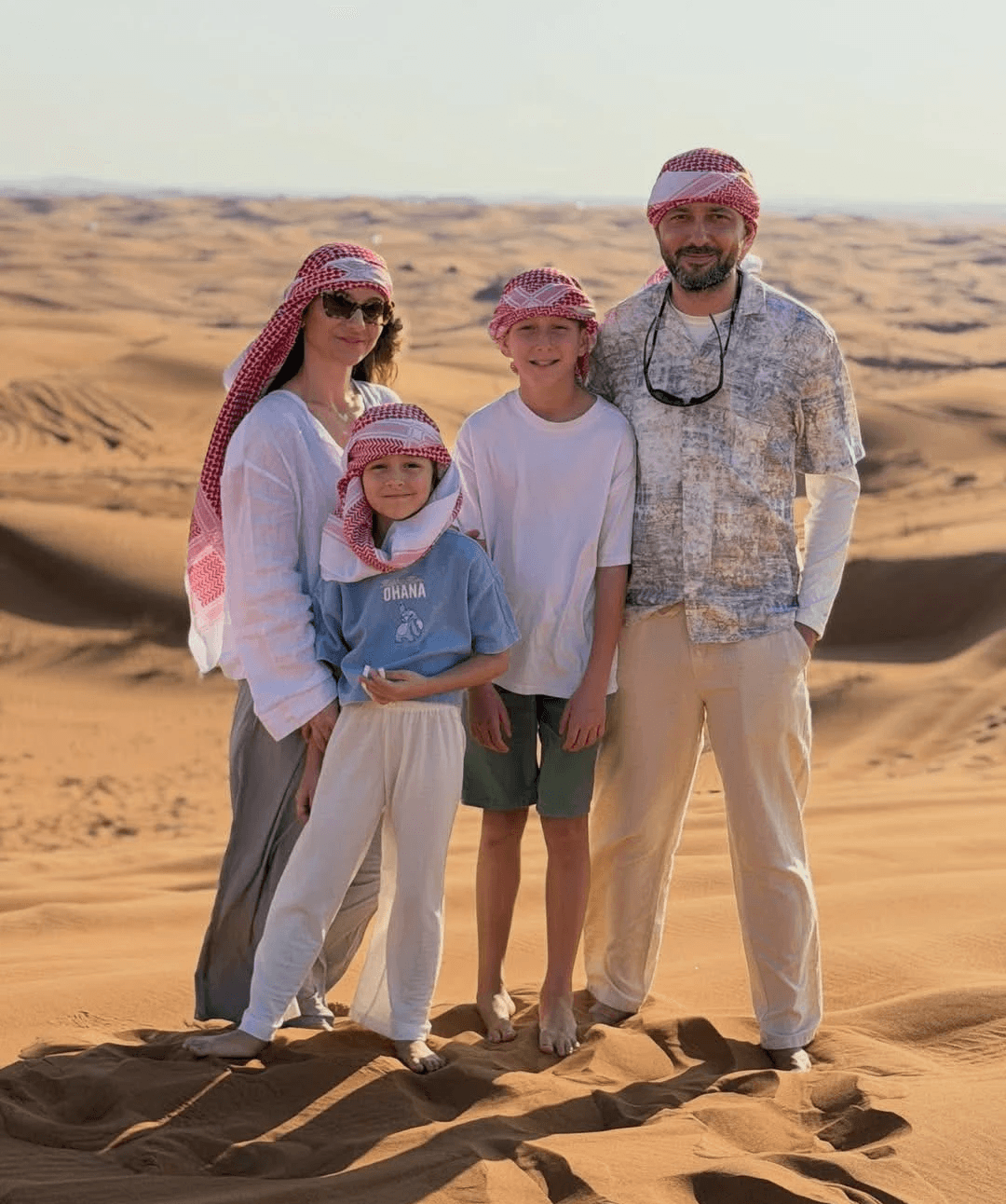 A family of four wearing traditional red and white keffiyehs, smiling together on the golden sand dunes during a private Dubai desert safari