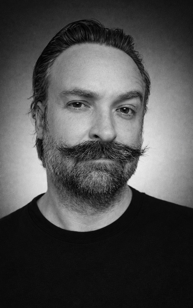 Woman smiling at computer desk with dual monitors in home office setup.