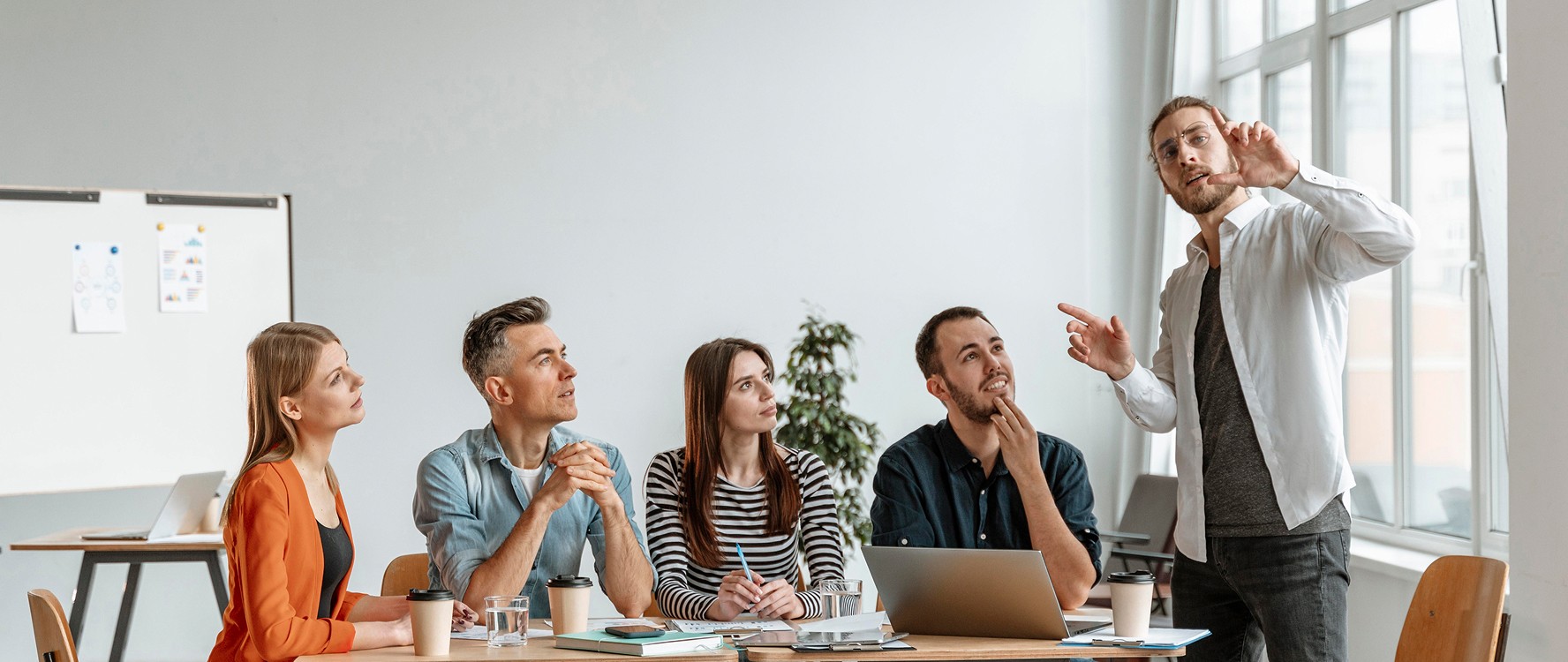 A group of five people sits around a wooden conference table in a bright office, attentively watching a standing presenter who gestures towards a whiteboard, surrounded by laptops and notebooks, emphasizing teamwork and collaboration in a professional meeting setting.