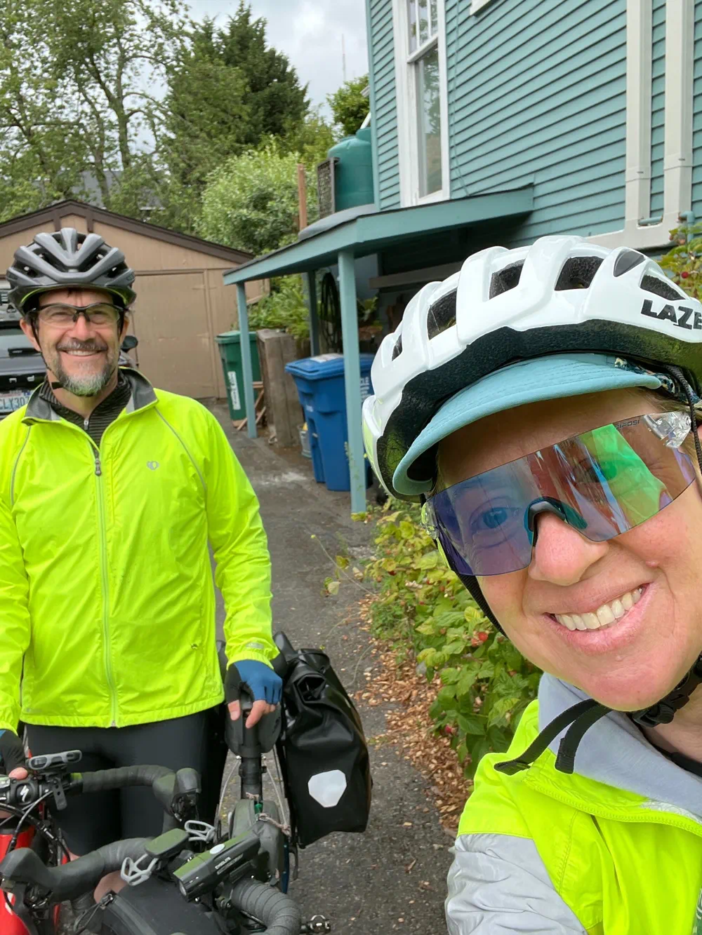 Two cyclists wearing helmets and bright rain jackets pause with their loaded bikes in a residential driveway before a ride.