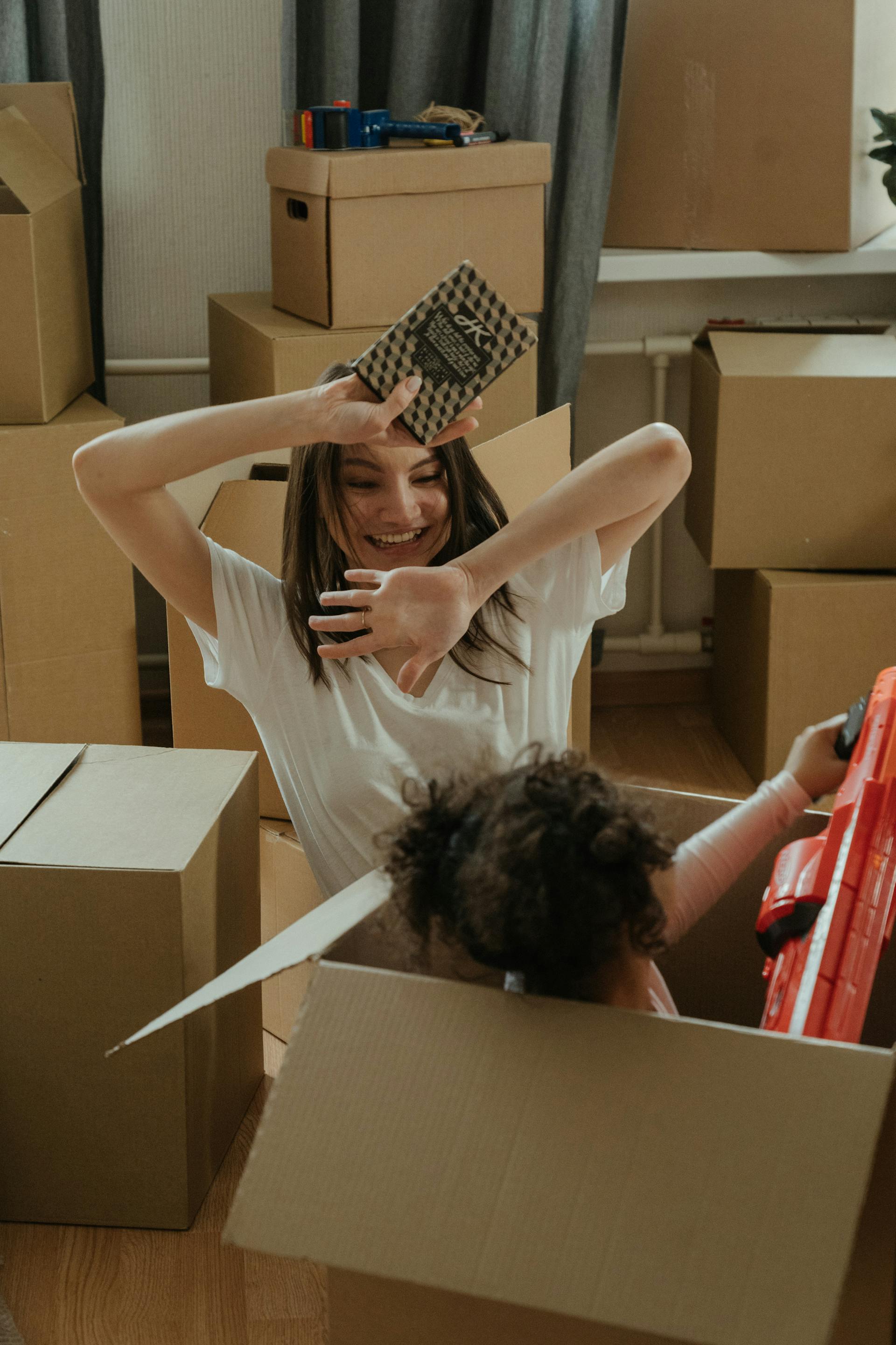 A young woman with long dark hair, wearing a white t-shirt, is smiling with her hands held over her head while sitting in a large cardboard box. A small child with curly dark hair is popping out of a box in the foreground, playing with a red toy gun. The room is surrounded by packed moving boxes.