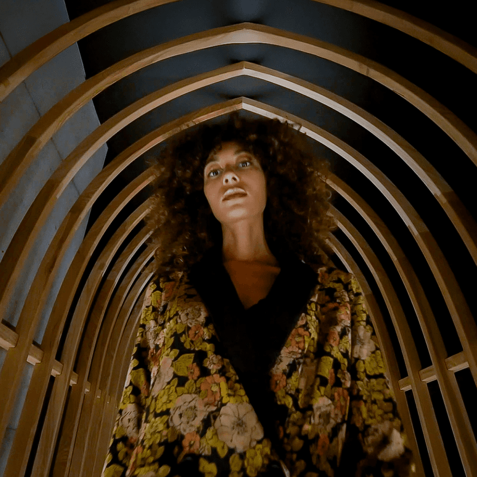 A woman with curly hair stands under a wooden arch structure, wearing a floral patterned jacket and looking straight ahead.
