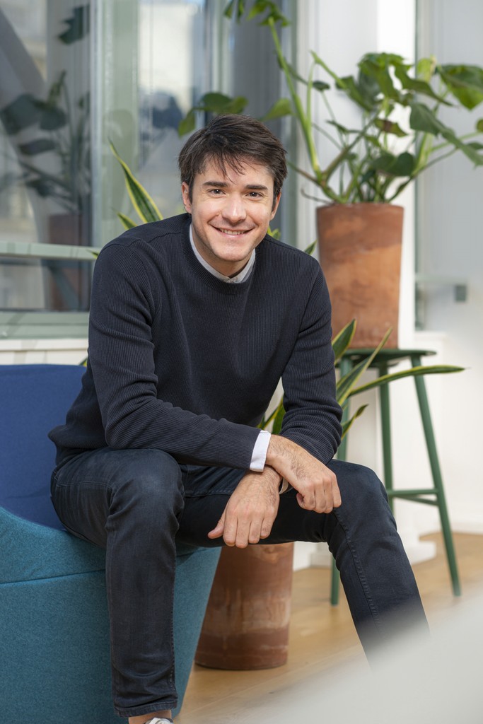 A young man with blond hair wearing a dark coat, a rust-colored vest, and a patterned tie, conveys a sophisticated and serious tone.