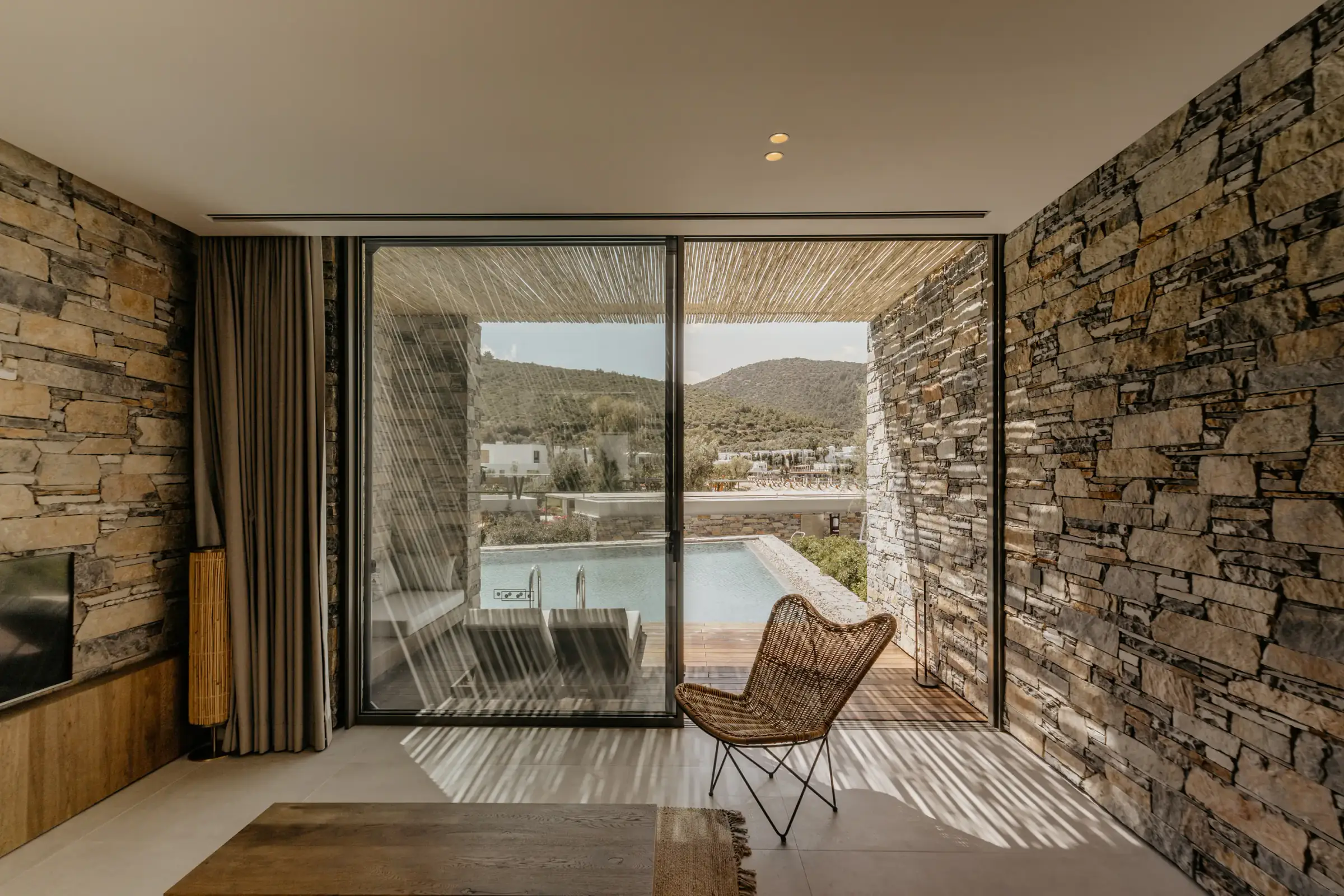 View from the room to the terrace and pool, with stone walls and a rattan chair