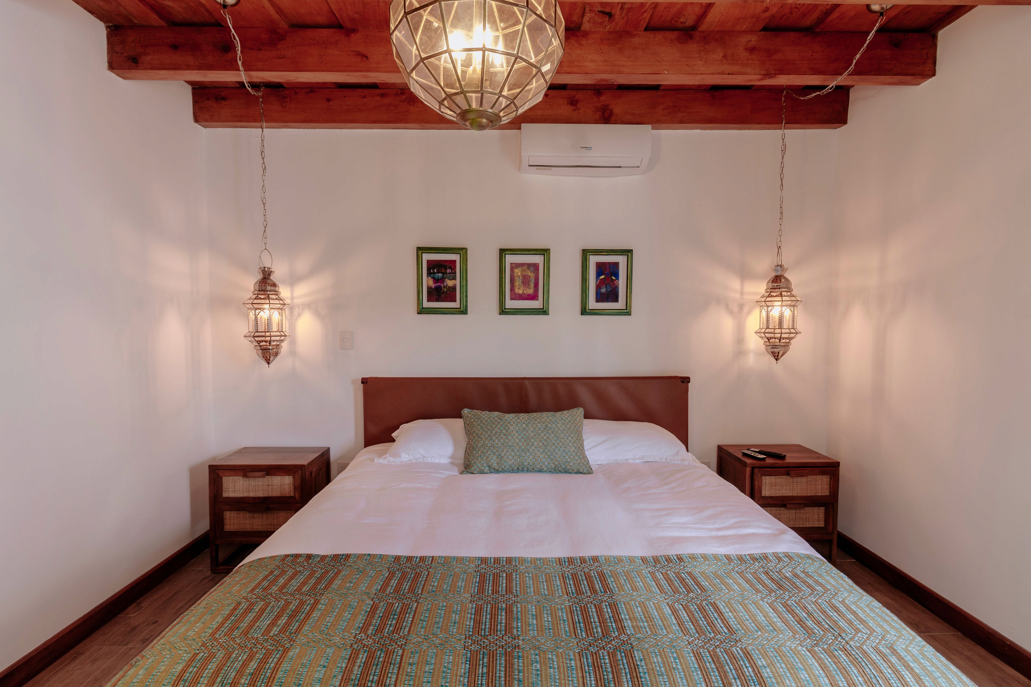 A cozy bedroom with a rustic wooden ceiling features a neatly made double bed adorned with a green and brown patterned throw, flanked by two wooden nightstands with hanging lantern-style lights, and three colorful framed artworks above the bed.