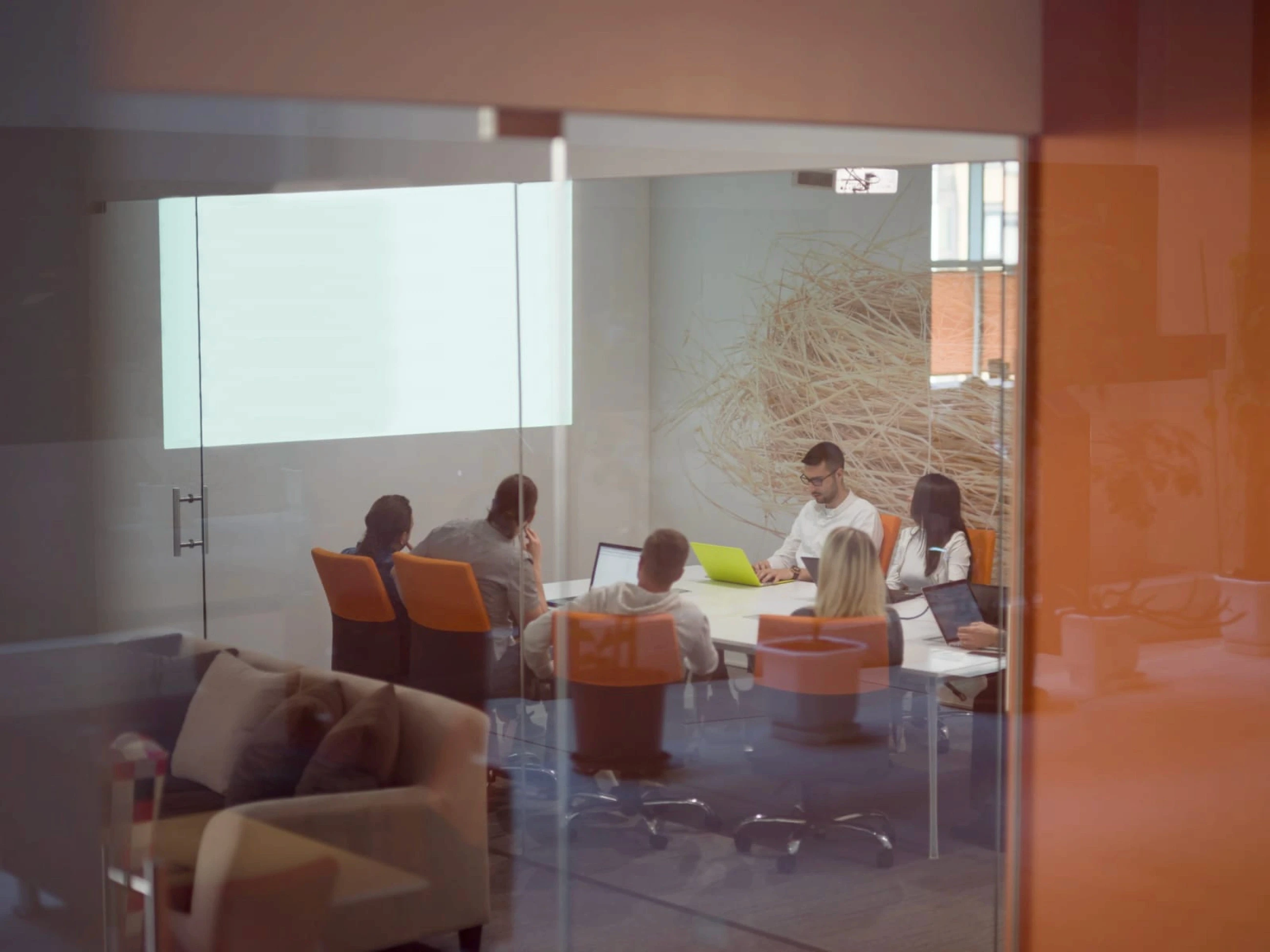 A group of people sit around a conference table in a modern office with a large window, engaged in a meeting while viewing a presentation on a wall-mounted screen.