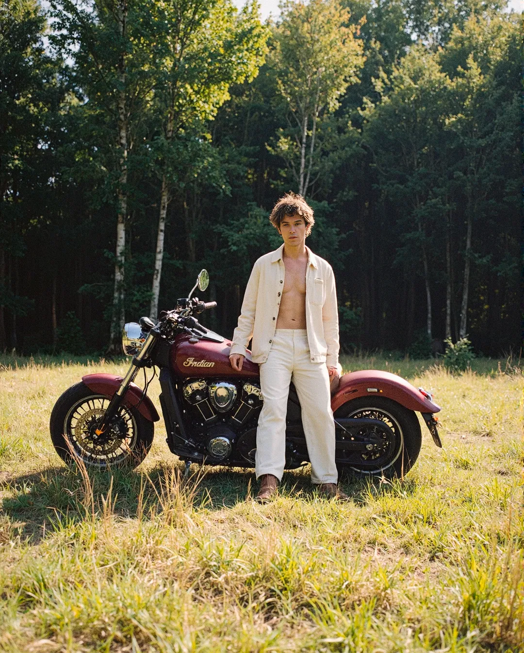Vintage-inspired summer fashion scene with a burgundy Indian motorcycle in a sunlit meadow bordered by green forest