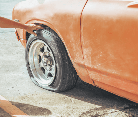 A close-up of a flat tire on an orange car. A hand is reaching out to touch the tire, which is visibly deflated. The car's body shows some dust and wear, and the background features a concrete surface, indicating an outdoor setting.