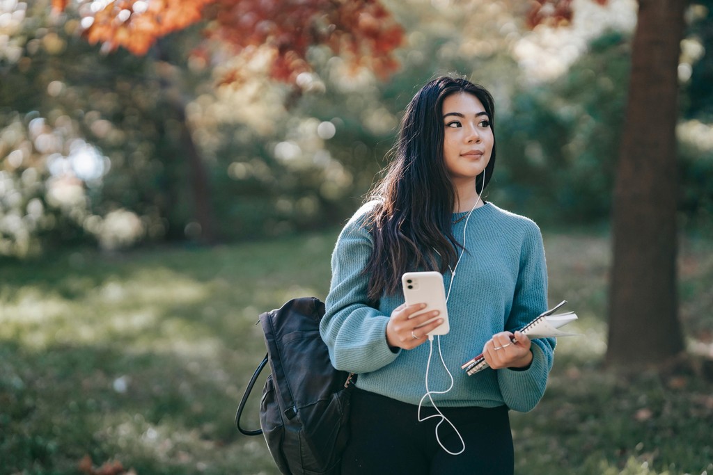 A female international student standing on a Canadian university campus in autumn, holding a smartphone and notebooks, representing newcomers searching for the best phone plan for students in Canada.