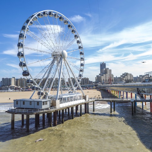 Una gran rueda de la fortuna en un muelle con vista a una playa de arena con edificios al fondo bajo un cielo azul.