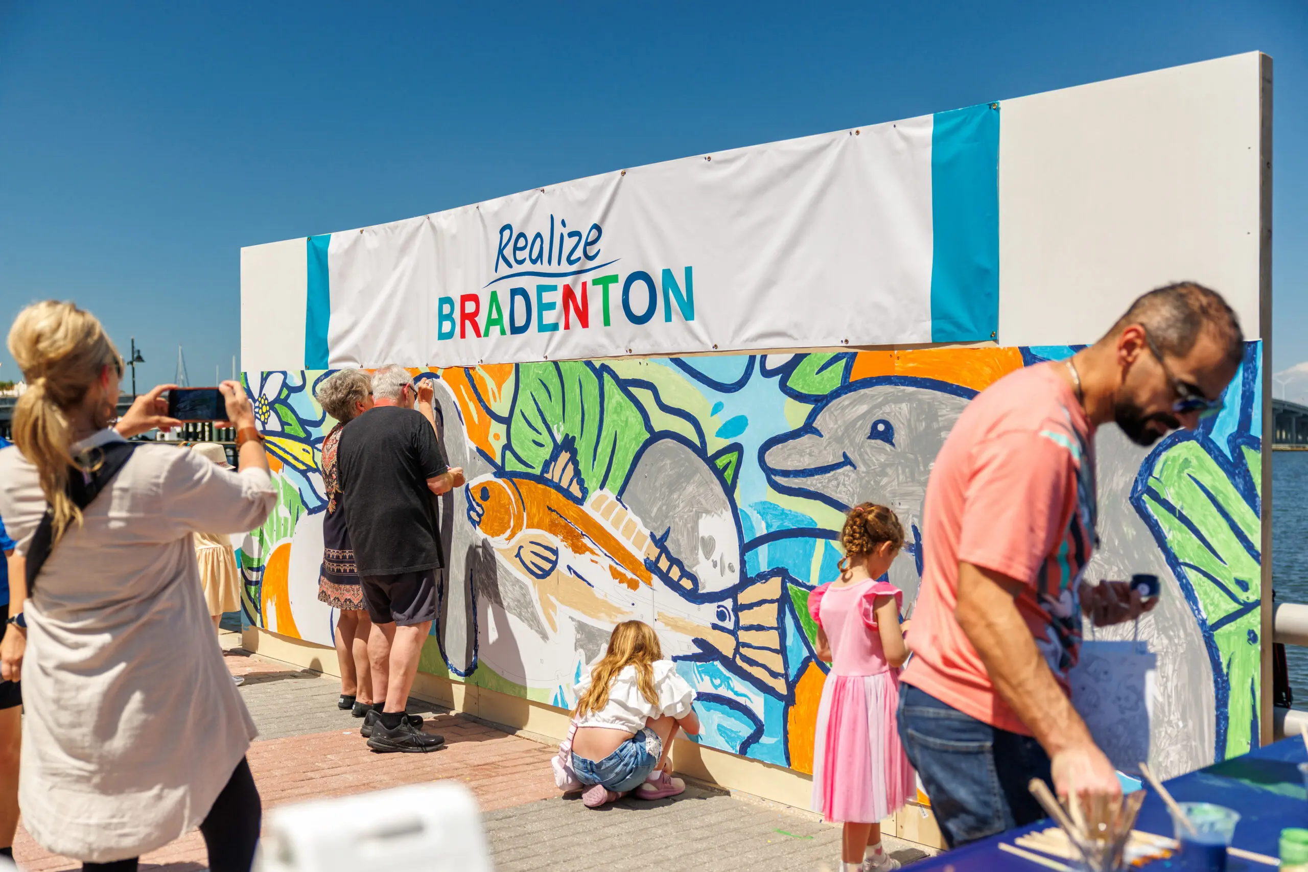 People engage in a collaborative mural painting under clear blue skies at the outdoor BAMFest event in Bradenton, FL, capturing the vibrant community spirit.