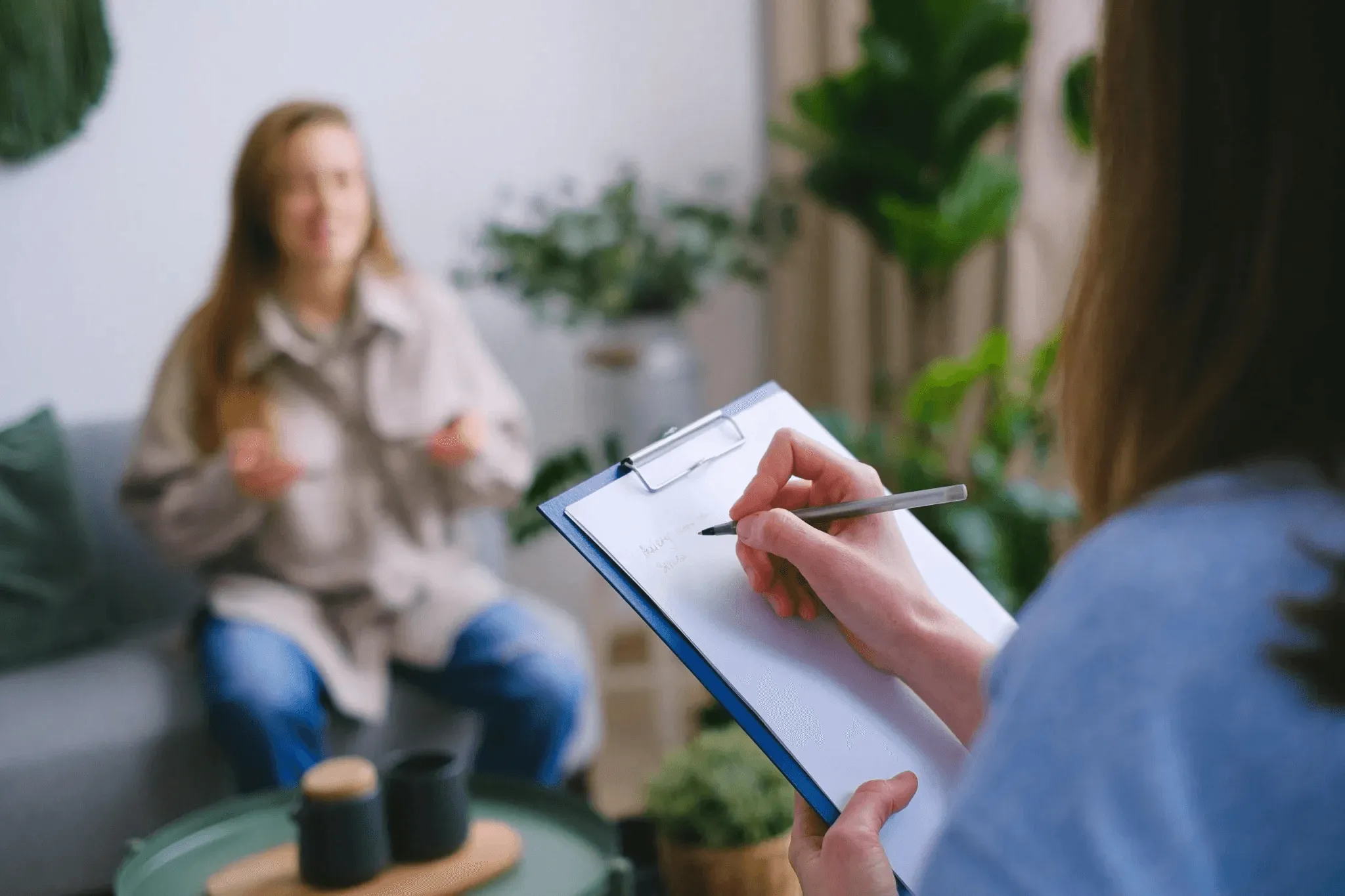 Therapist taking notes on a clipboard while a client speaks openly during a psychotherapy session