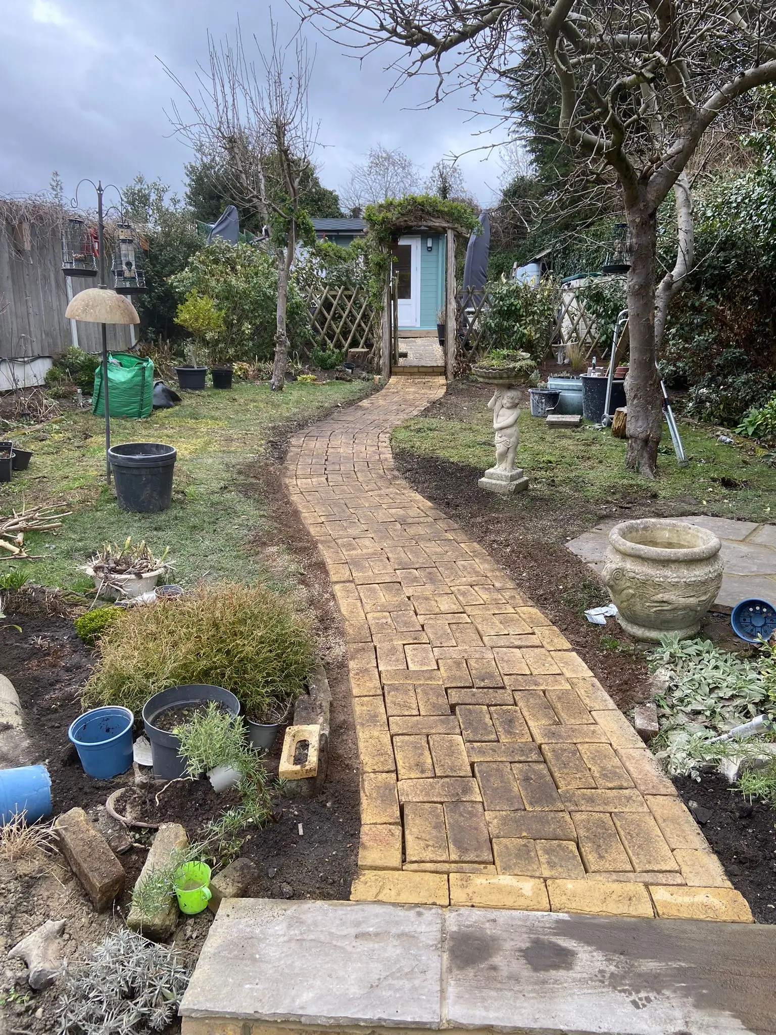 A winding garden path flanked by greenery and flower beds, leading towards a distant structure under a cloudy sky.