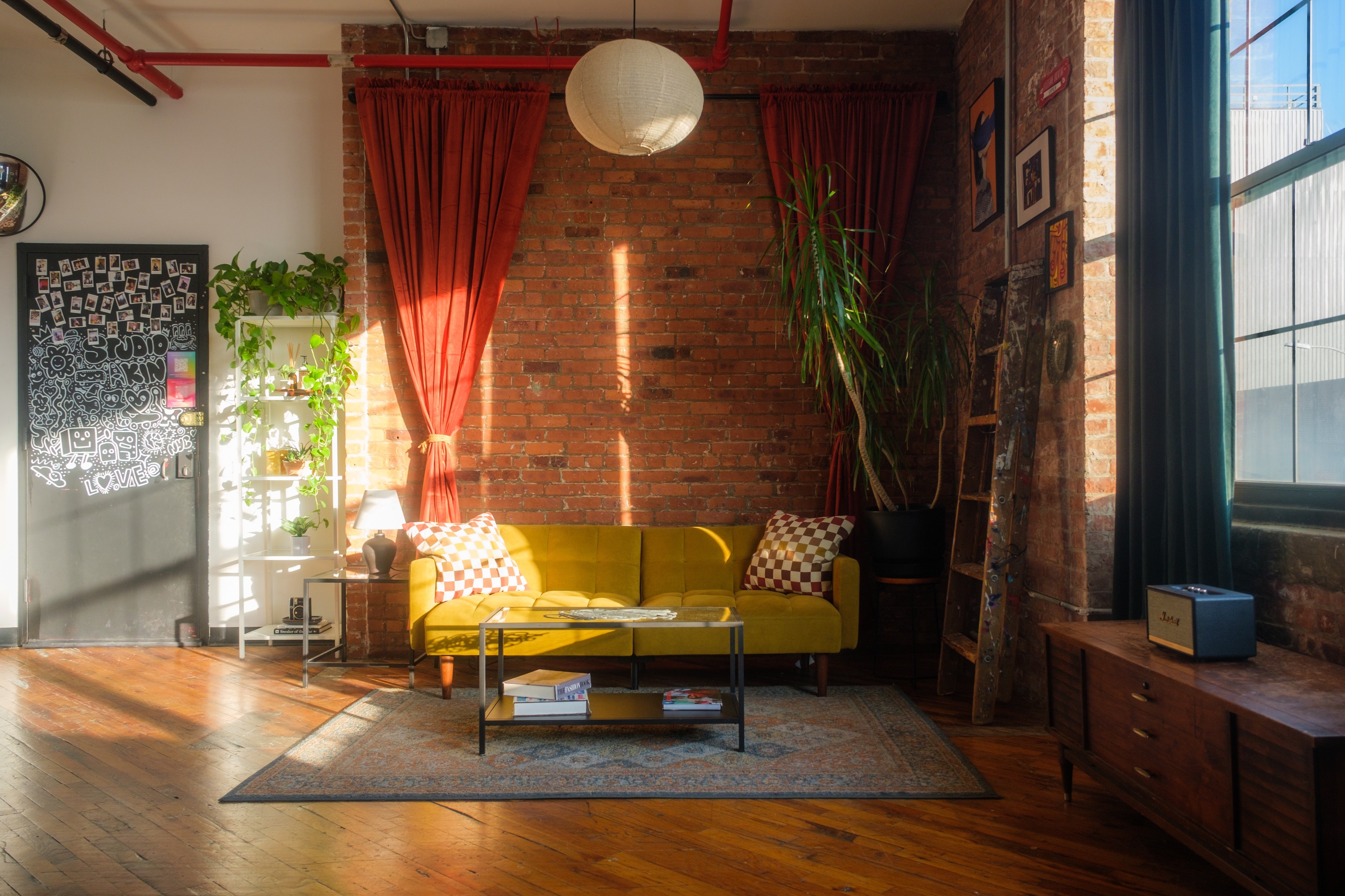 A sunlit room with exposed brick walls features a yellow couch with checkered pillows, vibrant plants, and eclectic decor, creating a cozy and inviting atmosphere. Natural light streams in from a large window with yellow curtains, adding warmth to the space.