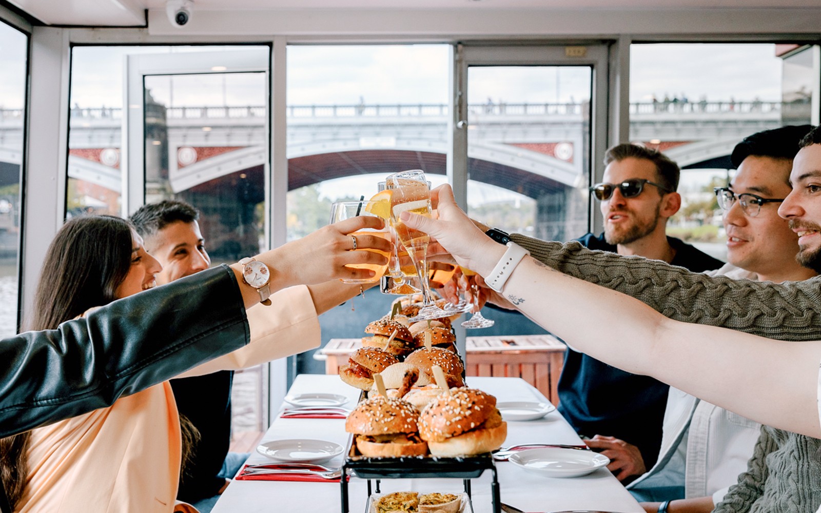 Guests toasting with drinks on a brunch cruise in Melbourne, with burgers on the table.