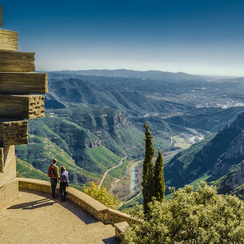 Two people stand on a balcony overlooking a vast valley with mountains, a river, and distant landscape, under a clear blue sky.