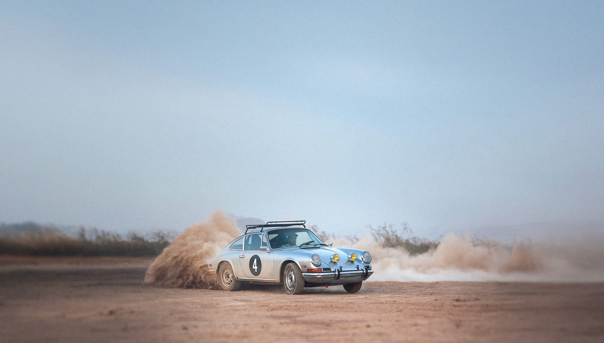 A vintage silver Porsche rally car with the number 4 on its side kicks up a cloud of dust as it drifts on a dirt track in a desert landscape.