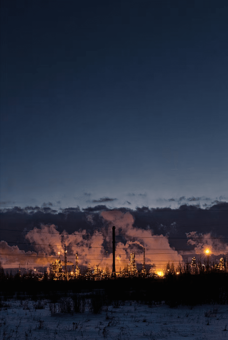 Industrial refinery with illuminated processing units and smoke stacks at dusk.