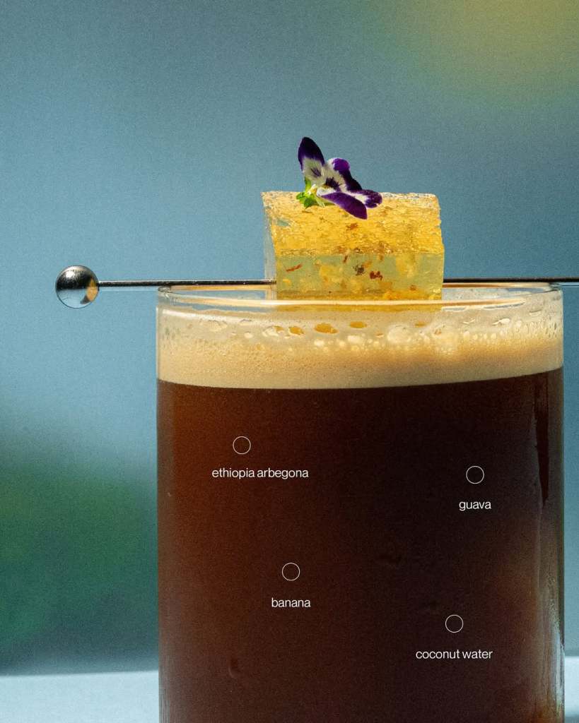 A close-up of a layered coffee drink topped with a clear jelly cube adorned with a flower, featuring labels for the flavors: Ethiopia Arbegona, guava, banana, and coconut water.