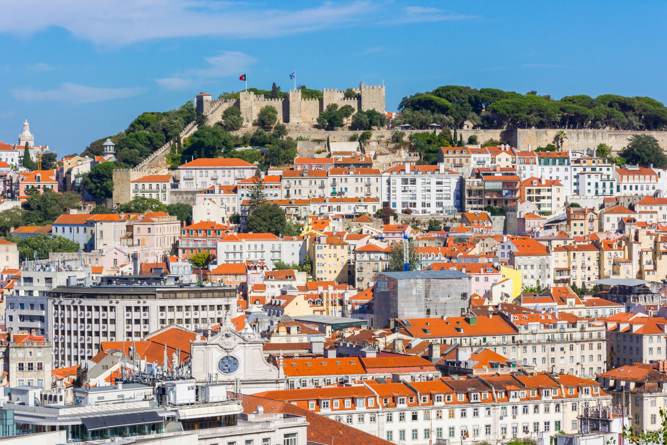A high-angle view of a coastal city with terracotta roofs and white buildings nestled against a hill topped by a stone