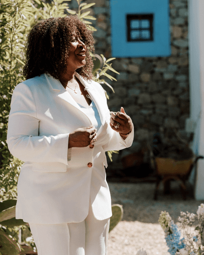 Zelfstandig trouwambtenaar Mildred Nijhove in een elegante witte outfit spreekt vol emotie tijdens een zonnige destination wedding in het buitenland.